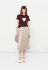 Mujer vistiendo una camiseta de logo Guess en burdeos, falda midi beige, sandalias blancas de deslizar, con un pequeño bolso de lavanda, de pie contra un fondo blanco.