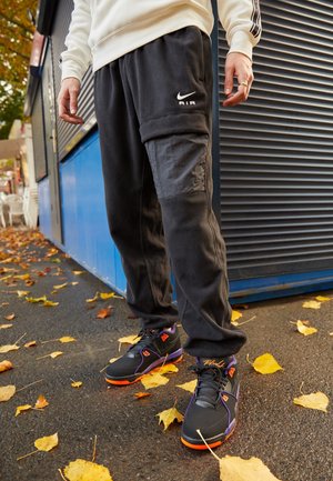 Person iført sorte Nike Air joggingbukser og sorte sneakers med lilla og orange accenter, stående på våd asfalt med efterårsblade.