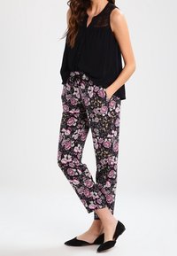Femme portant un haut sans manches noir avec des détails en dentelle, un pantalon à imprimé floral rose et blanc, et des ballerines noires à bouts pointus, debout les mains dans les poches.