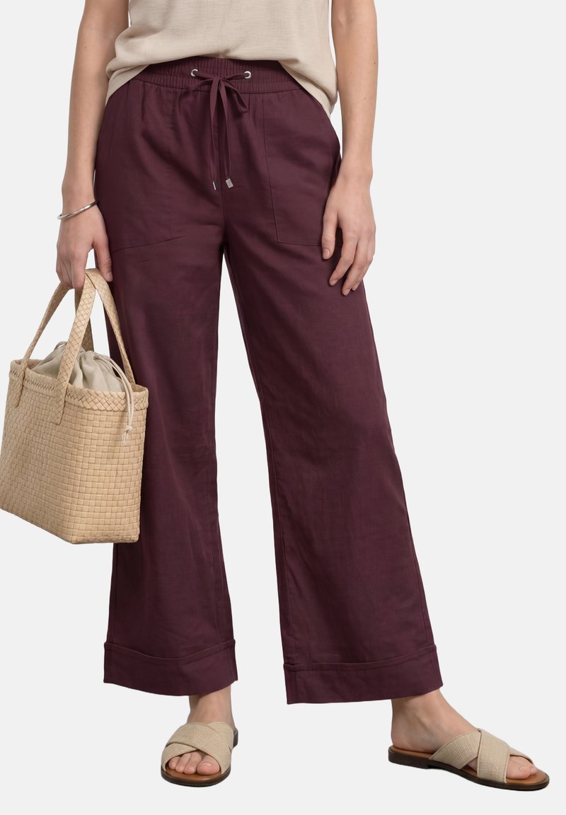 Femme portant un pantalon ample bordeaux avec une taille à cordon, tenant un sac à main beige tissé et portant des sandales beige à enfiler.