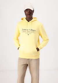 Gelber Hoodie mit einer Fronttasche, versehen mit einem Logo "Tommy Hilfiger" in Dunkelblau und Rot. Kombiniert mit beigen Hosen und einer hellen Kappe.