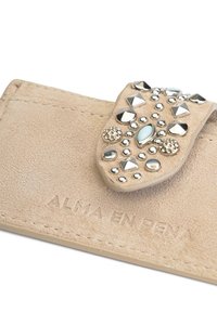 Cartera de ante beige con una tapa decorativa que presenta tachuelas metálicas y un acento en aguamarina. Logo grabado en la base.