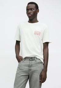 Tricou alb din bumbac cu "DAVID BOWIE" în litere portocalii și mov pe piept, asortat cu blugi din denim gri deschis.