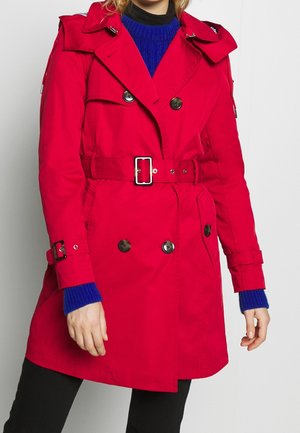 Kobieta nosząca jaskrawoczerwony trencz z paskiem, na niebieskim swetrze i czarnych spodniach, stojąca na jednolitym białym tle.