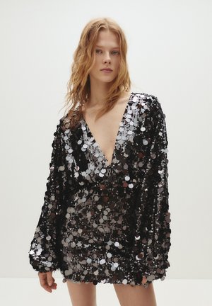 Robe noire à sequins avec un profond décolleté en V, des manches longues et un ourlet court. Les sequins sont circulaires et créent une surface réfléchissante.