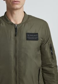 Olive green bomber jacket made from smooth, lightweight fabric. Features a zip front, ribbed collar, and a patch with "11 PROJECT" on the chest.