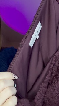 Vinrött tyg med mjuk textur. Innerfodret är slätt. Taggen med varumärkesnamn syns. En hand håller tyget med svarta naglar med prickar.