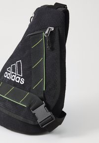Black fabric sling bag with textured surface, green accent lines, adjustable strap, and zippered pocket featuring an Adidas logo.