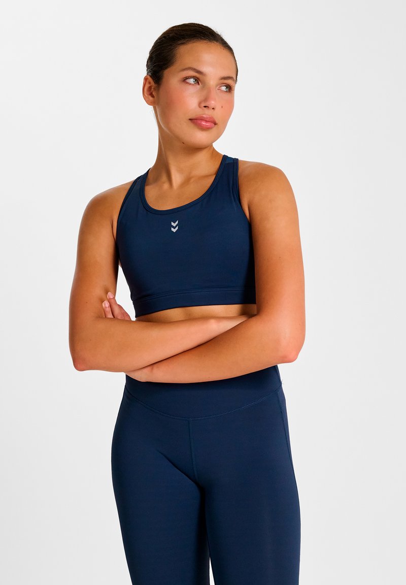 Marinblå sport-bh med slät yta och racerback-design. Har tre chevrondetaljer framtill. Matchas med stretchiga leggings i samma färg.