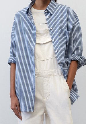 Osoba ubrana w białe ogrodniczki i niebiesko-białą pasiastą koszulę zapinaną na guziki, z podwiniętymi rękawami, na jednolitym tle.