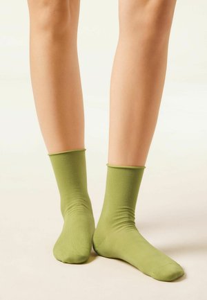 Legs wearing mid-calf length green socks standing on a light-colored surface with a plain light background.