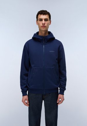 Homme portant un sweat à capuche bleu marine zippé avec poches avant et jean foncé, debout devant un fond clair uni.