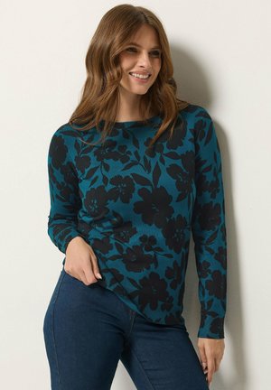Femme aux cheveux bruns portant un pull bleu canard avec des motifs floraux noirs et un jean bleu foncé, souriante et appuyée contre un mur blanc.