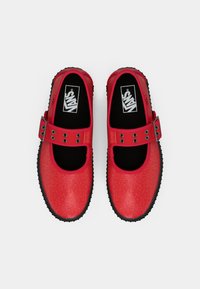 Rdeče bleščeče Mary Jane stil Vans čevlje z črnimi podplati in nastavljivimi zaponkami, pregledano od zgoraj na belem ozadju.