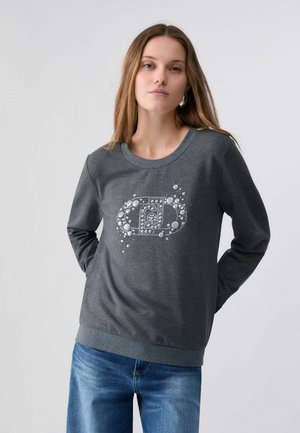 Frau mit langen Haaren trägt ein anthrazitgraues Sweatshirt mit einer perlenbesetzten Verzierung auf der Vorderseite und blaue Jeans, steht vor einem schlichten Hintergrund.
