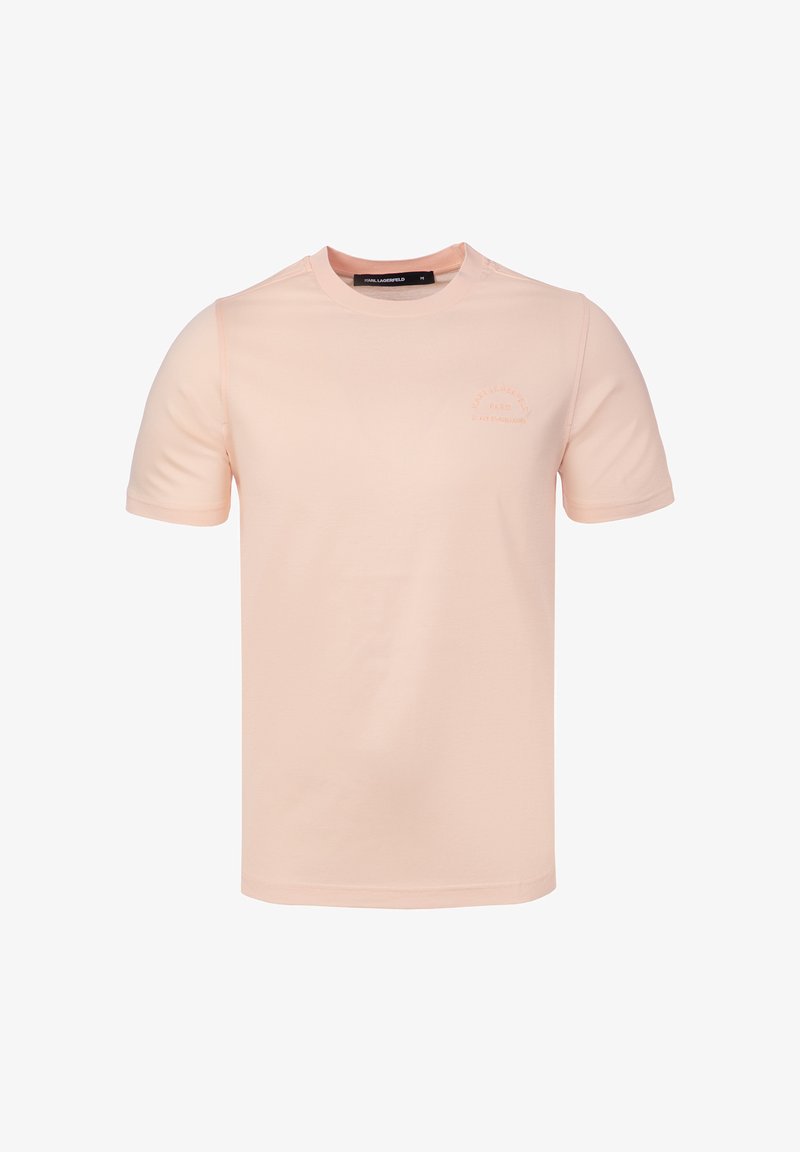 T-shirt rose clair à manches courtes et col rond avec un logo texte discret sur la poitrine gauche et une étiquette noire à l’intérieur du col, taille moyenne.