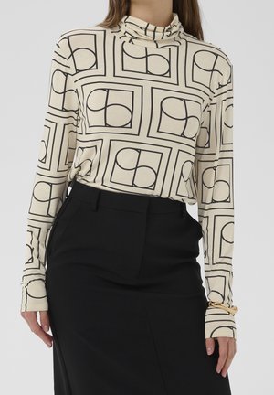 Femme portant un col roulé crème avec des motifs carrés géométriques noirs et une jupe noire taille haute, avec un bracelet doré au poignet.