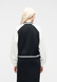 Άτομο με μακριά ξανθά μαλλιά που φοράει μαύρο και άσπρο varsity μπουφάν και ταιριαστή φούστα, με την πλάτη γυρισμένη σε απλό φόντο.