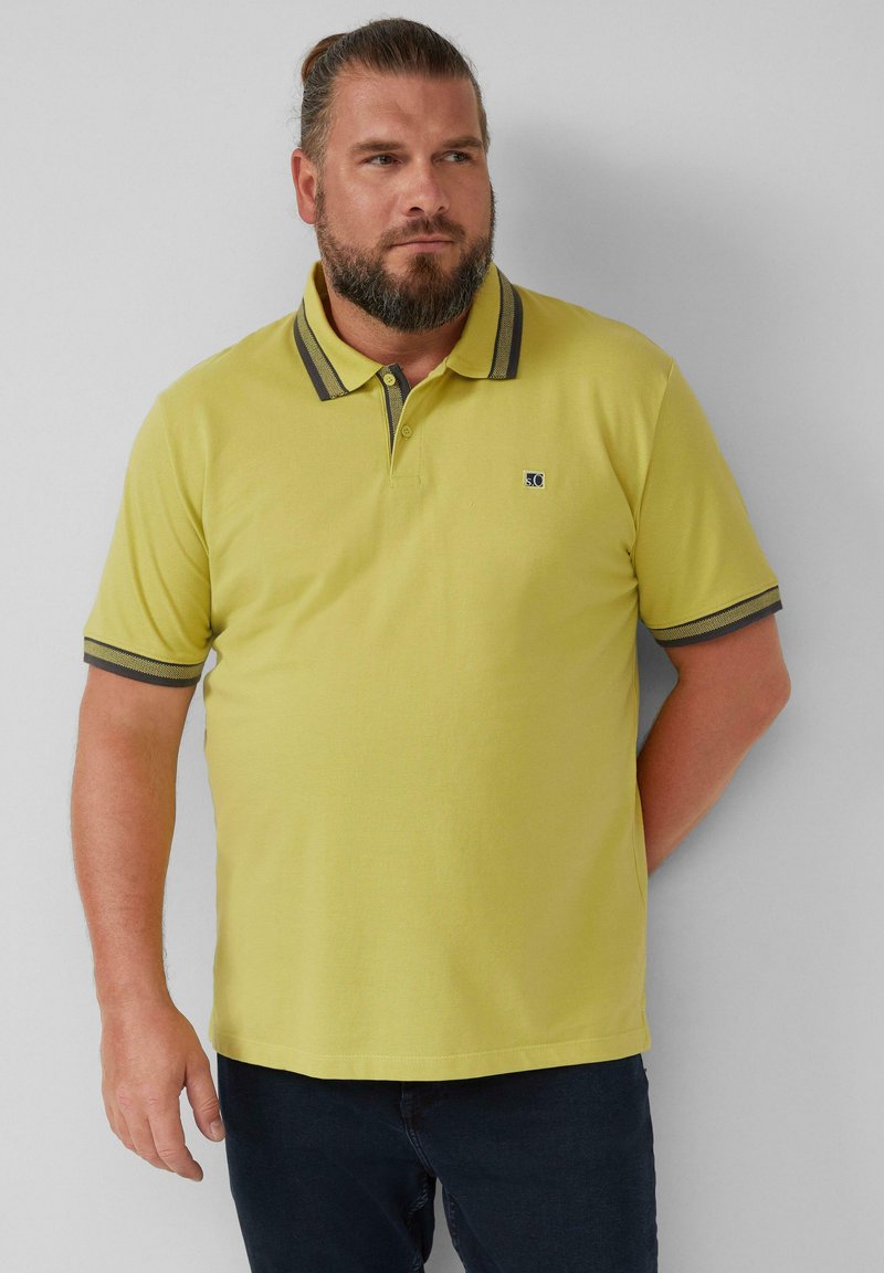 Gelbes Poloshirt aus strukturiertem Material, mit einem dunkel gestreiften Kragen und einem kleinen Logo auf der linken Brust, kombiniert mit einer dunklen Jeans.
