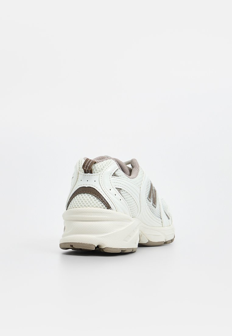 Zapatilla deportiva blanca con parte superior de malla texturizada y acentos en marrón claro. Tacón redondeado y suela acolchada diseñada para la comodidad y el soporte.