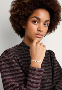 Sidabrinė apyrankė su kryžiaus pakabuku, uždėta ant švelnios grandinėlės su mažomis karoliukais. Modelis vilki tamsiai dryžuotą tekstūruotą marškinių viršų.