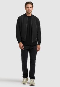 Jeune homme debout, portant une veste bomber noire, un jean noir, une chemise noire et des baskets beiges, sur un fond clair uni.