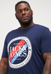 T-shirt corto blu navy in cotone, con un logo circolare che presenta "JACK & JONES" in bianco e contrasti rossi, colletto rotondo.