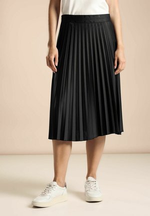 Vrouw draagt een zwarte midi-rok met plooien, gecombineerd met witte sneakers, staand tegen een effen beige achtergrond.