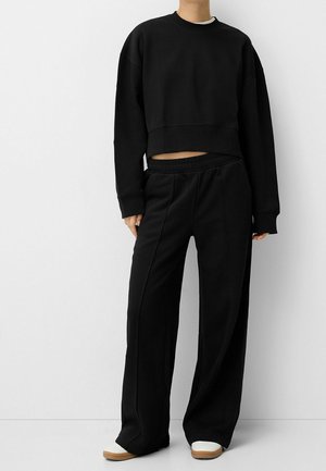 Persoon die een zwarte cropped trui met lange mouwen en losse zwarte broek met witte schoenen draagt, staand tegen een effen witte achtergrond.