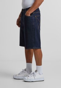 Shorts en denim foncé avec une coupe droite, poches latérales, accents de couture jaune et design à longueur de genoux, associés à des baskets blanches et des chaussettes montantes.