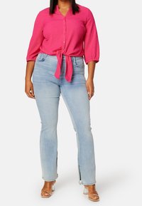 Blouse fuchsia à nouer devant avec manches trois-quarts, associée à un jean bleu clair à jambes droites avec fentes latérales aux chevilles.