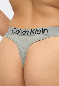 Γκρι βαμβακερό στρινγκ με λωρίδα λογότυπου που φέρει το "Calvin Klein" σε μαύρα γράμματα· απαλό ύφασμα με μινιμαλιστικό σχεδιασμό.