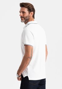 Vit pikétröja med ribbad krage som har en marinblå accent, korta ärmar och en vanlig passform, ihop med mörkblå jeans.