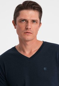 Granatowa koszulka z dekoltem w serek, wykonana z miękkiego materiału, z małym logo na lewej piersi, ma luźny krój i długie rękawy.