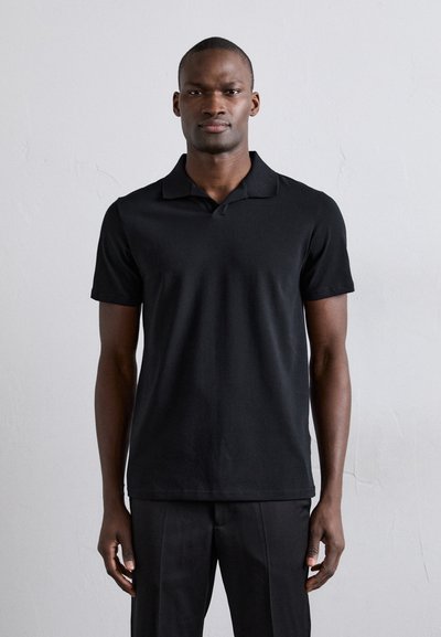Sort polo shirt lavet af glat stof; har krave, korte ærmer og lige snit. Båret med sorte bukser mod en lys baggrund.