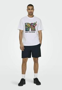 T-shirt bianco con grafica multicolore del logo MTV. Maniche corte e vestibilità comoda. Indossato con pantaloni corti blu navy e sneaker nere.