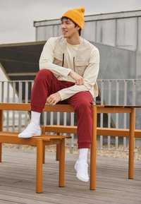 Homme portant un bonnet jaune, un gilet beige, une chemise crème et un pantalon rouge, assis sur un banc orange en plein air, un pied posé sur un banc inférieur.