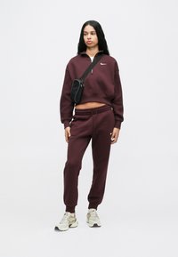 Donna che indossa un completino Nike maroon con un top corto con zip, pantaloni jogger abbinati, sneaker beige e una borsa a tracolla nera, in piedi su uno sfondo bianco.