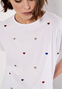 Hvid bomulds t-shirt med spredte broderede hjerter i rød, blå og guld, med en afslappet pasform og korte ærmer.