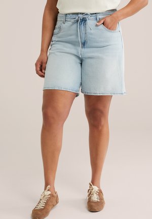 Jasnoniebieskie krótkie spodenki dżinsowe z regulowanym sznurkiem w pasie, noszone z beżowymi sneakersami z sznurowadłami, na stojącej osobie.