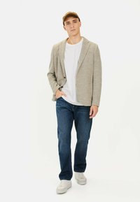 Helles graues strukturiertes Blazer über einem weißen T-Shirt, kombiniert mit blauen Jeans und weißen Sneakern. Eine Mütze in einem gedämpften Braunton.