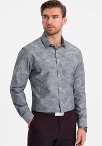 Camisa abotonada de tela gris con un estampado floral, mangas largas, cuello extendido y un bolsillo en el pecho. Combinada con pantalones oscuros.