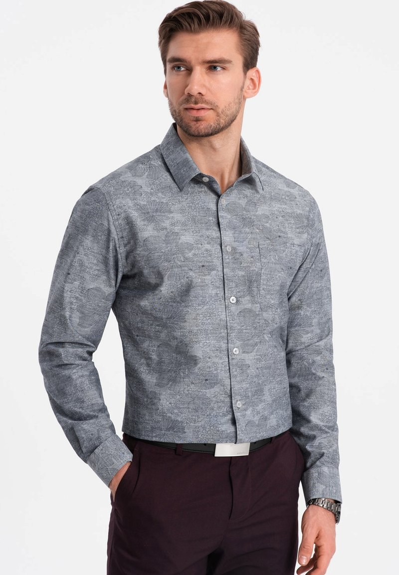 Camisa abotonada de tela gris con un estampado floral, mangas largas, cuello extendido y un bolsillo en el pecho. Combinada con pantalones oscuros.