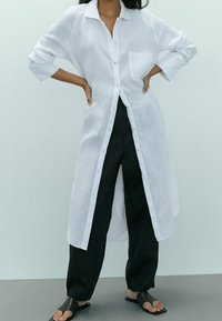 Vrouw in een lange witte knoopshirt over zwarte wijde broek en zwarte open tenen sandalen, staand met handen op de heupen tegen een effen achtergrond.