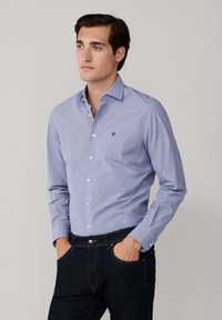 Giovane uomo con capelli scuri che indossa una camicia azzurra chiara con bottoni e jeans scuri, in piedi con le mani in tasca su uno sfondo neutro.