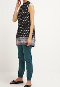 Czarna tunika z geometrycznymi białymi wzorami i kolorową lamówką, zestawiona z dopasowanymi turkusowymi spodniami. Modelka ma na sobie płaskie czarne sandały.