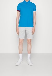 Blauwe poloshirt met rode en zwarte gestreepte accenten, gecombineerd met lichtgrijze shorts en witte sneakers. Model staat tegen een effen achtergrond.