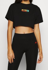 Černé zkrácené tričko s krátkými rukávy a barevným logo "ellesse". Nosené s odpovídajícími černými legínami s vysokým pasem.