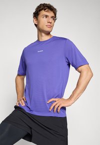 Ung mand iført en lilla Salomon T-shirt og sorte shorts står med hænderne på hoften og kigger mod venstre foran en ensfarvet baggrund.