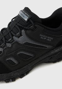 Juodai Skechers Slip-Ins, su sintetine viršutine dalimi, tekstūrinėmis medžiagomis, guminėmis padais ir atspindinčiais akcentais matomumui.